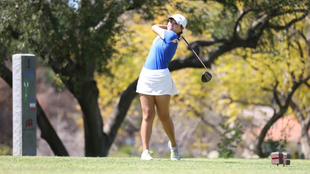 Katelyn Vo makes a stroke on the golf course. Vo wears the golf uniform of a white cap and white skirt with a blue shirt.